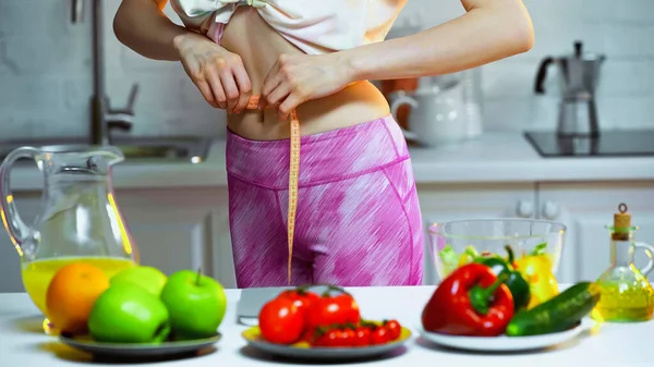 Cropped view of woman measuring waist near vegetables and fruits on blurred foreground — Stock Photo