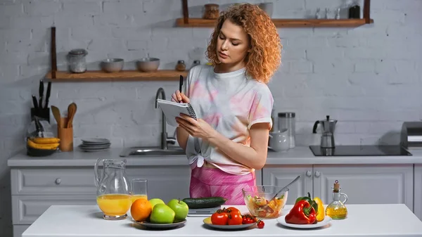 Mulher encaracolada escrevendo em caderno e pesando pepino perto de legumes e suco de laranja na mesa — Fotografia de Stock