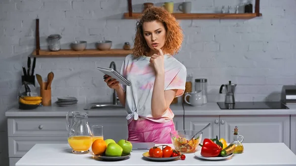 Nachdenkliche Frau hält Notizbuch neben Gemüse und Obst auf Küchentisch — Stockfoto