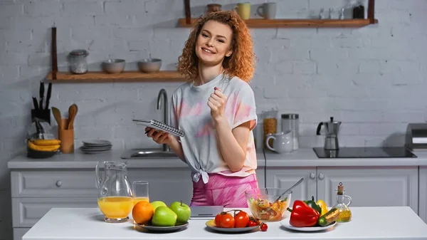 Mulher sorrindo segurando notebook perto de legumes e frutas na mesa da cozinha — Fotografia de Stock
