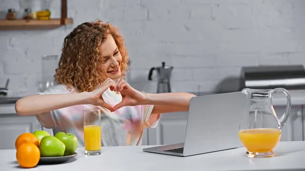 Кудрявая молодая женщина смотрит на ноутбук, показывая знак сердца с руками во время видеозвонка — стоковое фото