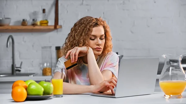 Кучерява молода жінка тримає кредитну картку під час покупки в Інтернеті на кухні — стокове фото