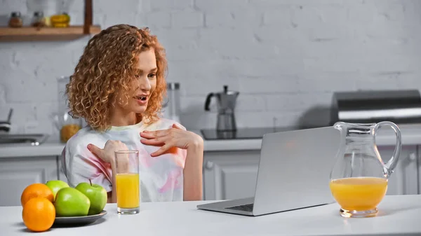 Кучерява молода жінка вказує на себе під час розмови під час відеодзвінка на кухні — стокове фото