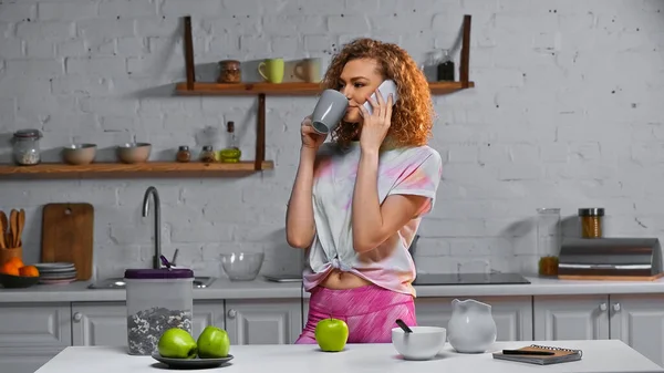 Кучерява жінка говорить на смартфоні і п'є чай біля кукурудзяних пластівців і яблук на столі — стокове фото