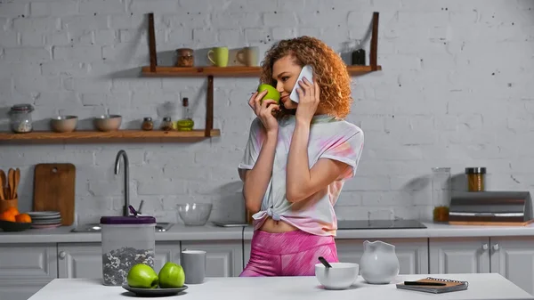 Curly woman talking on smartphone and smelling apple near corn flakes in container on table — Stock Photo