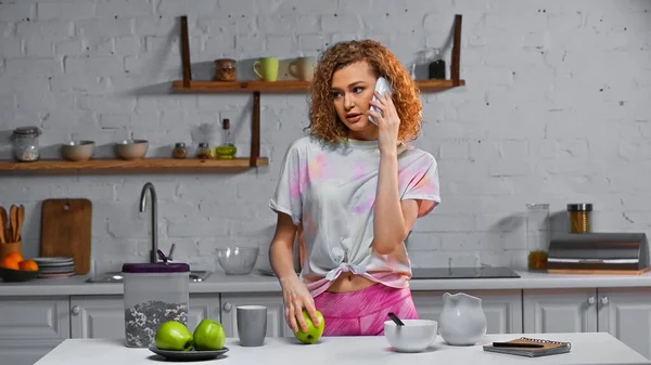 Curly woman talking on smartphone and holding apple near corn flakes in container on table — Stock Photo