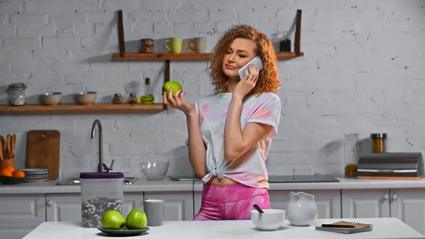 Curly young woman talking on smartphone and holding apple near corn flakes in container on table — Stock Photo