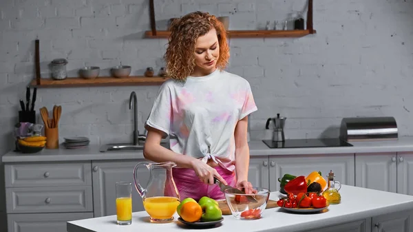 Frisé jeune femme coupe tomate fraîche dans la cuisine — Photo de stock