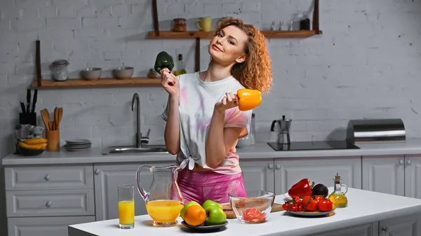 Curly young woman holding bell peppers near fresh veggies in kitchen — Stock Photo