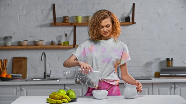 Feliz jovem adicionando flocos de milho na tigela perto de frutas na mesa da cozinha — Fotografia de Stock