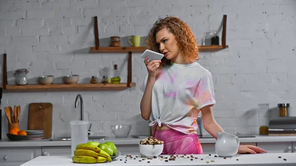 Curly woman recording voice message near corn flakes and fruits on kitchen table — Stock Photo