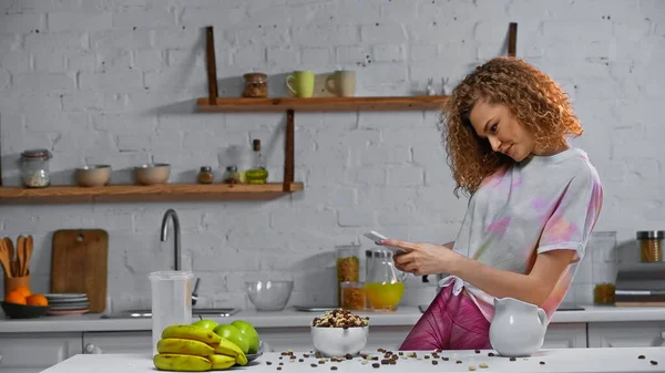 Mujer rizada tomando fotos de hojuelas de maíz y frutas en la mesa de la cocina - foto de stock