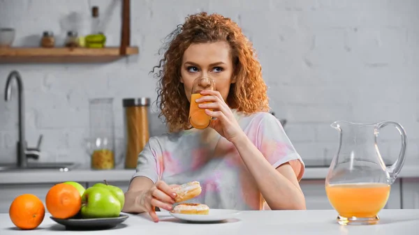 Femme bouclée tenant beignet et boire du jus d'orange dans la cuisine — Photo de stock