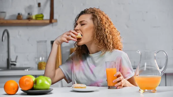 Femme bouclée manger beignet sucré et tenant un verre de jus d'orange dans la cuisine — Photo de stock