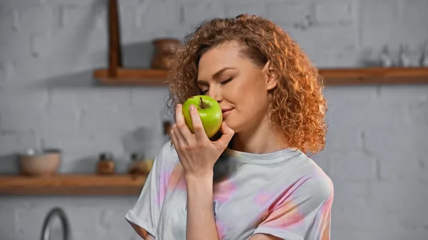 Curly young woman smelling ripe apple in kitchen — Stock Photo