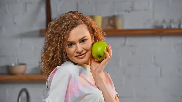 Веселая и кудрявая женщина держит спелое яблоко на кухне — стоковое фото