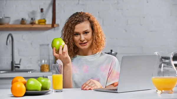 Curly woman holding apple while looking at camera near laptop and fruits on table — Stock Photo