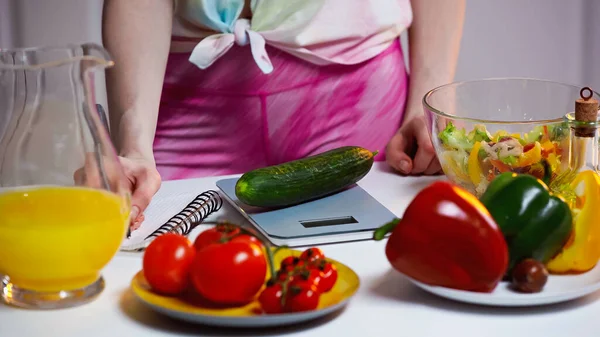 Cropped view of woman writing in notebook while weighing cucumber — Stock Photo