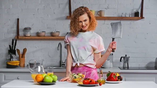 Donna allegra che tiene il coltello enorme vicino a insalata fresca in boccia e frutta su tavolo — Foto stock