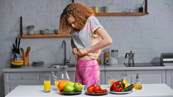 Curly woman measuring waist with measuring tape near veggies on kitchen table — Stock Photo