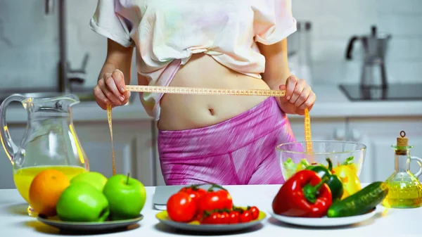 Partial view of woman measuring waist near vegetables and fruits on blurred foreground — Stock Photo