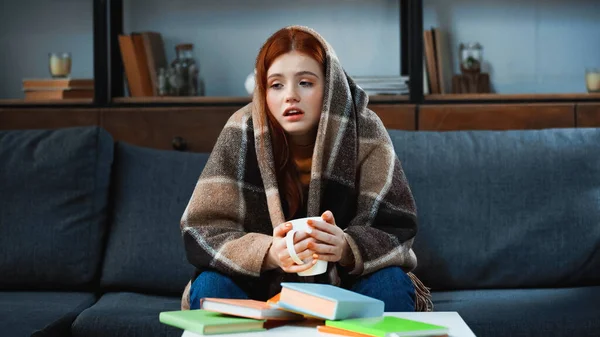 Red haired girl wrapped in blanket holding cup near books on blurred foreground — Stock Photo