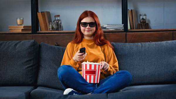 Smiling girl in 3d glasses holding popcorn and remote controller at home — Stock Photo