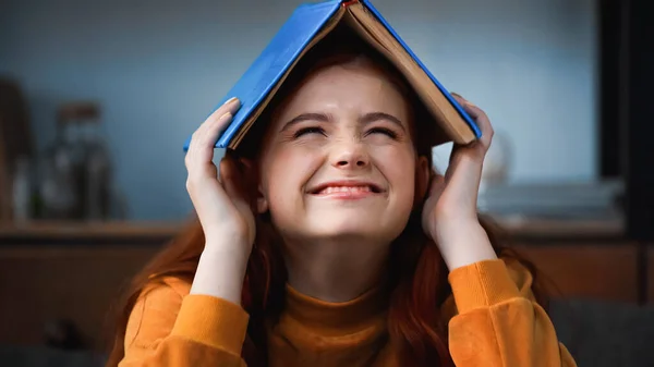 Feliz adolescente sosteniendo libro por encima de la cabeza en casa - foto de stock