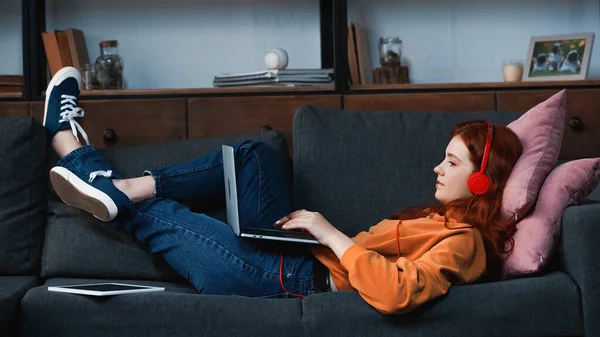 Side view of girl in headphones using laptop near digital tablet on couch — Stock Photo