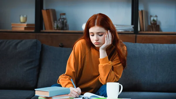 Estudiante molesto escribiendo en un cuaderno cerca de la taza y libros - foto de stock