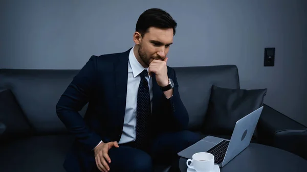 Uomo d'affari pensieroso seduto vicino al laptop e alla tazza di caffè in hotel — Foto stock