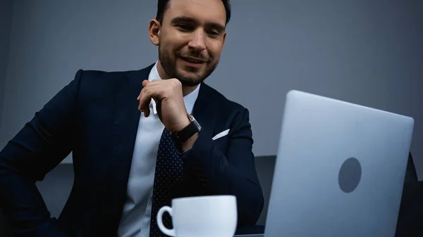 Smiling businessman looking at laptop near cup on blurred foreground in hotel — Stock Photo