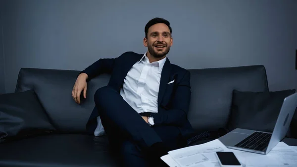 Smiling businessman looking away near gadgets and papers on blurred foreground in hotel — Stock Photo