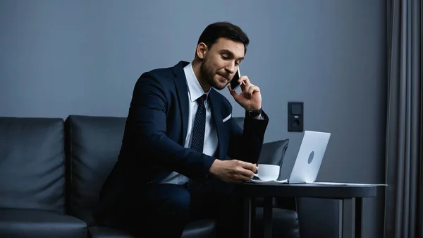 Empresário falando no smartphone e segurando xícara de café perto de laptop no hotel — Fotografia de Stock