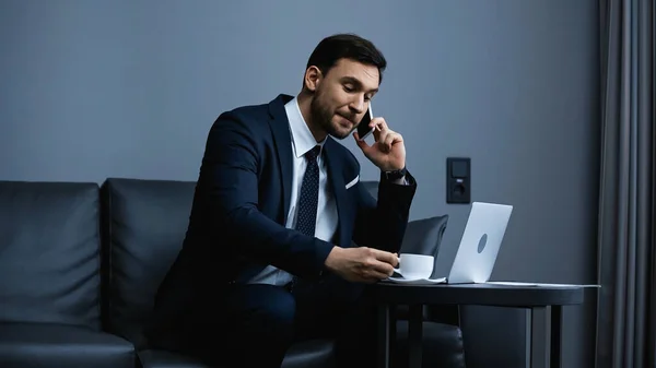 Empresario hablando por teléfono inteligente y sosteniendo la taza de café cerca de la computadora portátil en el hotel - foto de stock