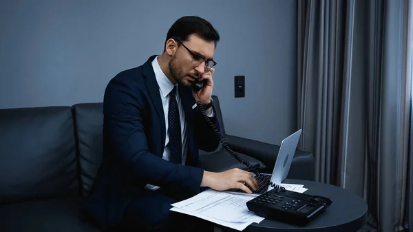 Менеджер в костюме разговаривает по телефону возле ноутбука и документов в отеле — стоковое фото