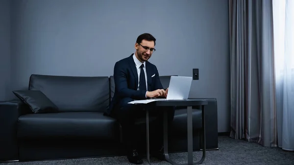 Uomo d'affari sorridente che digita su computer portatile vicino a documenti sul tavolo in camera d'albergo — Foto stock