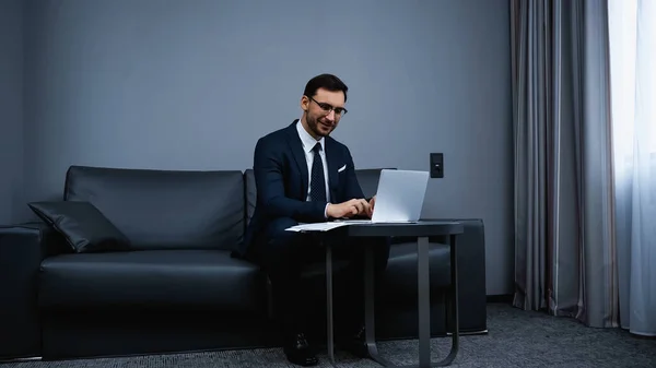 Empresário sorridente digitando no laptop perto de documentos na mesa no quarto de hotel — Fotografia de Stock