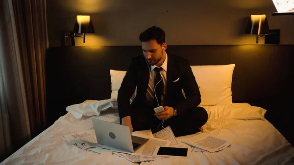 Businessman in earphones using laptop while holding smartphone near papers on hotel bed — Stock Photo