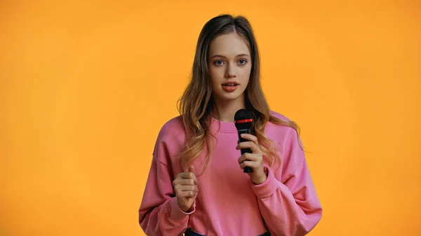 Teenage girl in pink sweatshirt talking while holding microphone isolated on yellow — Stock Photo