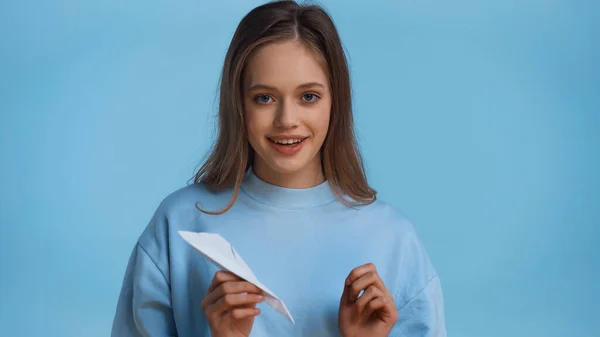 Happy teenage girl in sweatshirt holding paper plane isolated on blue — Stock Photo