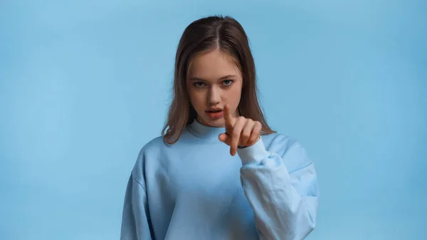 Дівчина-підліток в сорочці вказує пальцем, дивлячись на камеру ізольовано на синьому — стокове фото