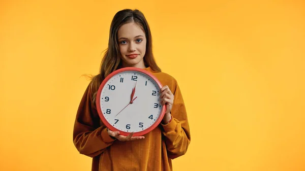 Souriant adolescent fille en pull tenant horloge isolé sur jaune — Photo de stock