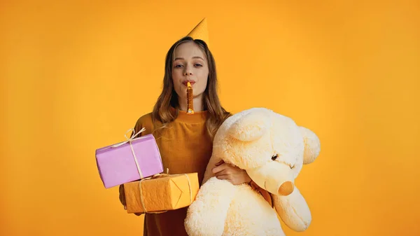 Teenager-Mädchen in Partymütze bläst Party-Horn, während sie Geschenke und Stofftier auf gelb isoliert hält — Stockfoto