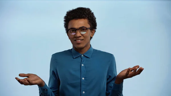 Jeune adolescent afro-américain en lunettes montrant un geste d'haussement d'épaules isolé sur bleu — Photo de stock
