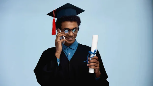 African american teenager in graduation gown and cap holding diploma while talking on smartphone isolated on blue — Stock Photo