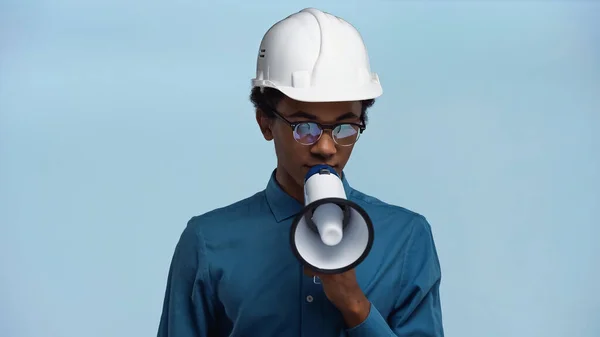 African american teenage boy in hard hat holding megaphone while talking isolated on blue — Stock Photo