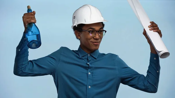 Adolescente afroamericano en sombrero duro sosteniendo rollo de papel y botella con champán aislado en azul - foto de stock