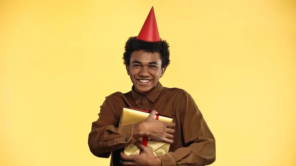 Alegre afroamericano adolescente en partido gorra celebración presente aislado en amarillo - foto de stock