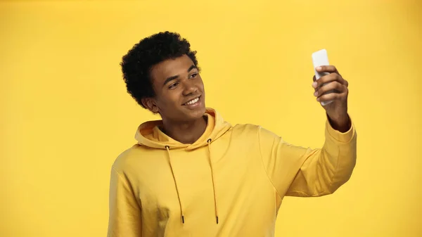 Happy african american teenage boy in hoodie taking selfie isolated on yellow — Stock Photo